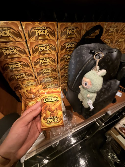 A hand holds a gold-colored "Gods Legends: Rise of the Pack" trading card pack in front of a display of identical packs, with a backpack featuring a green plush keychain hanging nearby.