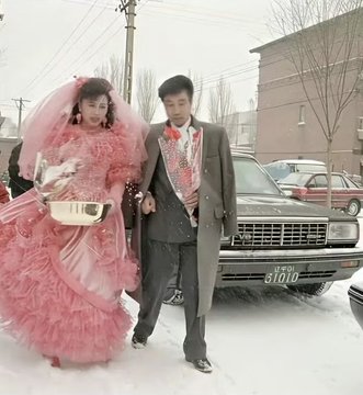 A bride in a voluminous pink dress and a groom in a grey suit walk together in a snowy setting, standing in front of a parked pickup truck.