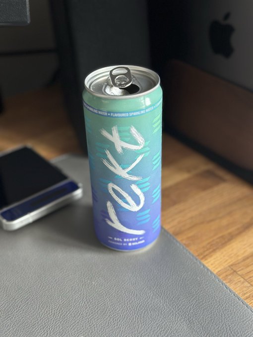 The image features a can of soda, possibly Rekt, placed on a gray surface next to a cell phone. The text "Boom they did it again! @rektdrinks absolutely smashing it out the park with @solana berry" is written above and below the soda can. This suggests that someone has posted about the soda brand or flavor on social media, possibly tagging the company or sharing their thoughts on the product.