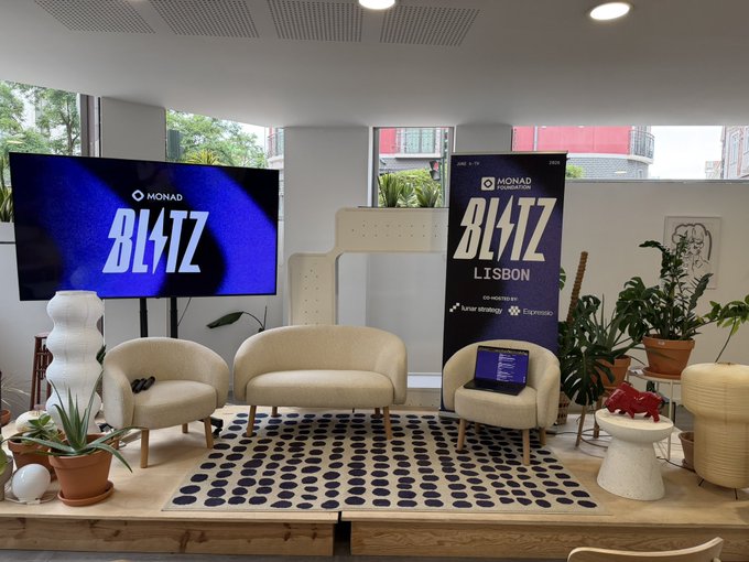 The image shows an event taking place inside a room, featuring two TVs displaying the word "Blitz" along with a banner that says "Lisbon". The room has white furniture, including chairs and couches arranged around a large rug on which there are potted plants placed. A laptop is also present in the scene. The main focus of the image is the event taking place within this space, likely an AI hackathon or similar gathering where people come together to work on innovative projects and share ideas.