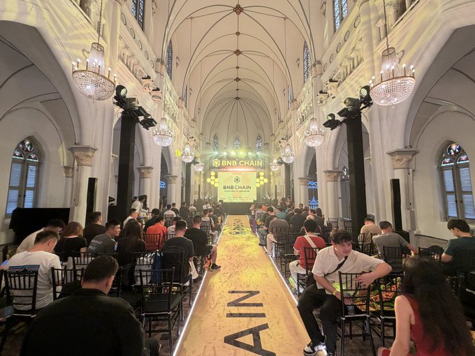 A fashion show runway event for 'BNB CHAIN' takes place in an ornate hall featuring vaulted ceilings and chandeliers, with attendees seated on both sides of a yellow runway displaying partial text.