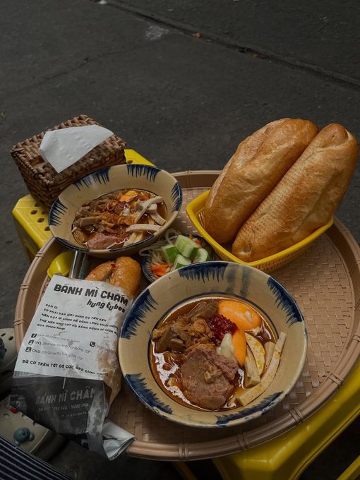 A top-down view of a woven tray holding a Vietnamese meal, including a bowl of noodle soup with sliced pork, fried spring rolls, and baguettes, alongside a napkin printed with 'BÁNH MÌ CHẢM'.