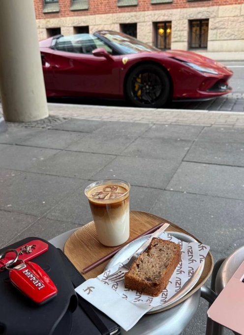 A glass of iced latte and a slice of banana bread on a tray at a cafe, with a red Ferrari parked on the street in the background, and a red Ferrari keychain visible on the table.