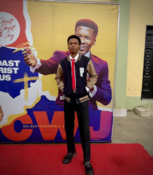 A young man wearing a varsity jacket with a 'P' patch, white shirt, and red tie posing on a red carpet in front of a banner featuring 'Christ In Joy' ministry artwork and the text 'SURPASSING'.