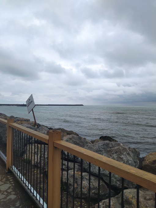 A coastal view features a wooden railing and large rocks along a stone path, with a tilted white sign overlooking choppy gray ocean water under a cloudy overcast sky.