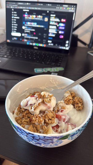 A bowl of yogurt with granola and fruit sits on a desk in front of a laptop displaying Discord, with a 'GRIP' sticker on the laptop.