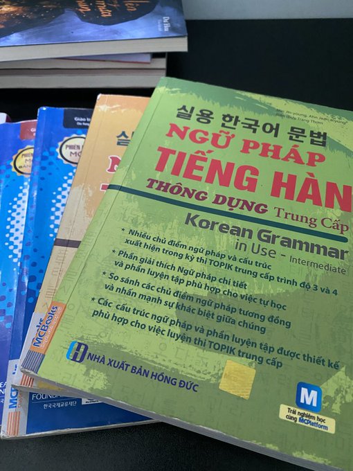 A photograph of stacked books with the top book prominently displayed, titled 'NGỮ PHÁP TIẾNG HÀN THÔNG DỤNG Trung Cấp' (Korean Grammar in Use - Intermediate) published by Nhà Xuất Bản Hồng Đức, featuring text in both Korean and Vietnamese.