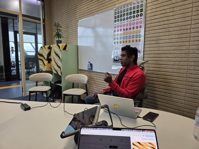 A man in a red jacket sits at a conference table with a laptop and tablet, gesturing while speaking in a room with a whiteboard displaying educational charts and a number line.