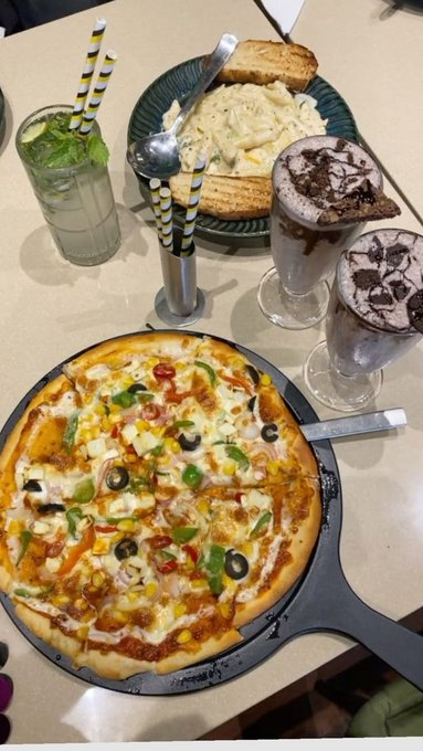 A meal spread featuring a vegetable-topped pizza on a black pan, a plate of pasta with toast, two chocolate milkshakes topped with cookies, and a refreshing drink with mint and black-and-white striped straws.