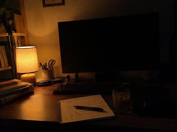 A dimly lit desk setup featuring a monitor, a glowing table lamp, a notebook with handwritten notes, and a glass of water.