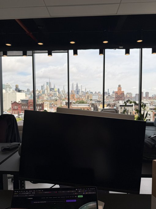 A dark computer monitor and keyboard sit in an office with floor-to-ceiling windows overlooking the Manhattan, New York City skyline under a partly cloudy sky.