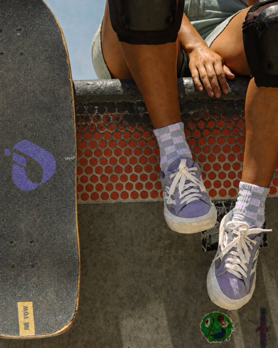 A skateboarder wearing purple sneakers and lavender-striped socks sits beside a skateboard featuring a purple logo, capturing the everyday street culture aesthetic that aligns with InterLink's message of becoming woven into daily life. The brand "WOW" is visible on the skateboard's grip tape, and a frog sticker rests on the ground.