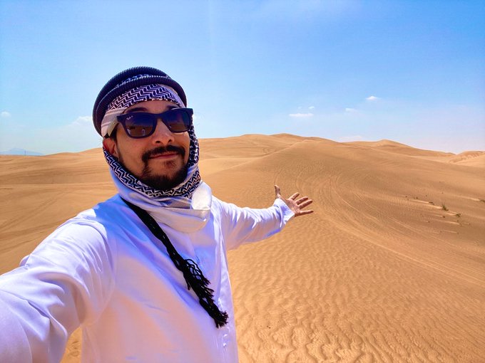 A selfie of a man wearing a white thawb, keffiyeh, and sunglasses, standing in a vast orange sand desert with rolling dunes under a blue sky.