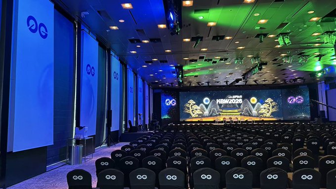 An empty auditorium set up for a conference, with rows of chairs and screens displaying the 'HBW2026' logo and a circular 'OG' emblem.