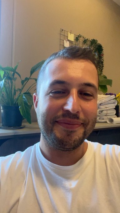A smiling man with a beard takes a selfie indoors, with potted plants and folded towels visible in the background.