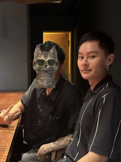 Two young men pose at a dimly lit restaurant table, with the person on the left playfully hiding their face behind a cartoon cat sticker wearing sunglasses, fitting the post's humorous question about why "墨山猫" (Ink Mountain Cat) won't reveal their face.