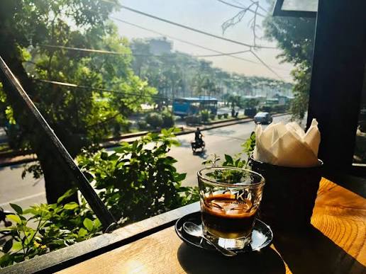 A view from inside a café showing a glass of iced coffee on a wooden counter, with a view through the window of a street scene with motorbikes and lush green trees in daylight.