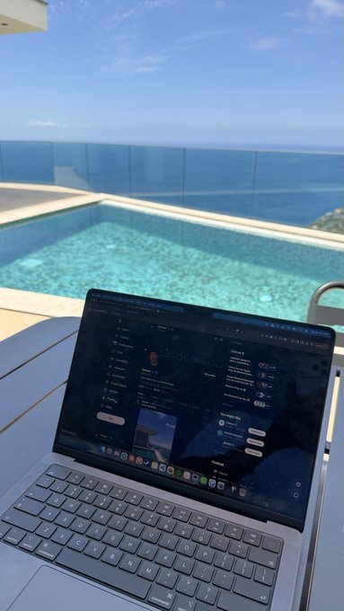 A MacBook sits open on a patio overlooking a stunning ocean view and infinity pool, displaying what appears to be an X (formerly Twitter) feed. The post invites others to share their Solana ($SOL) wallet addresses, suggesting the user is requesting crypto payment details while enjoying a luxurious morning setting.