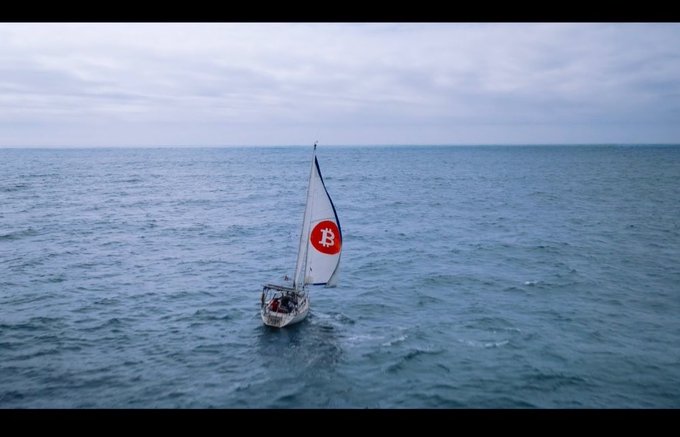An aerial view of a small sailboat with a Bitcoin logo on its sail, sailing on a calm ocean under a cloudy sky.