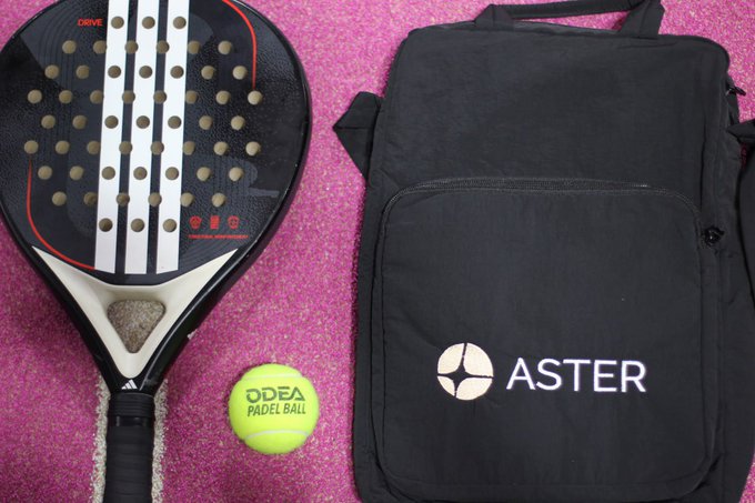 A black and white padel racket and an ODEEA padel ball sit next to a black bag with 'ASTER' branding on a pink textured surface.