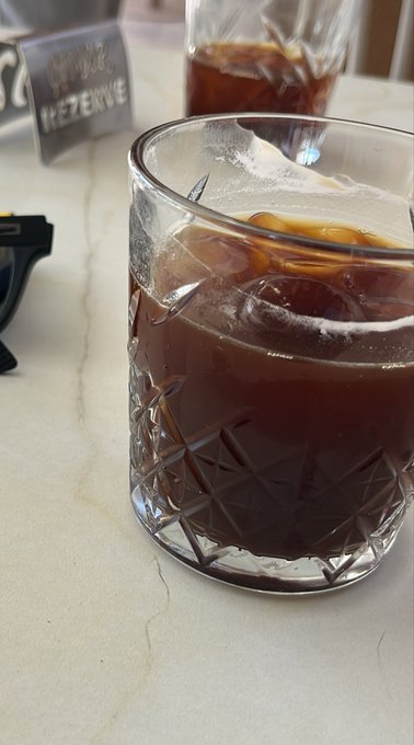 A close-up photo of a crystal-cut glass filled with a dark iced beverage, likely cold brew coffee, sitting on a white marble table near a 'RESERVE' sign.