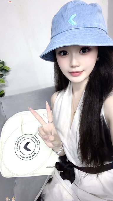 A selfie of a young woman wearing a light blue bucket hat and white top, giving a peace sign while holding a Michael Kors handbag with a visible logo.