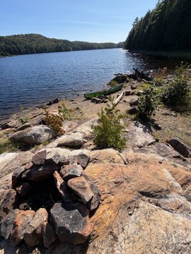 A scenic photograph of a rocky lakeshore with a stone fire ring in the foreground, calm water, and a forested horizon under a clear blue sky.