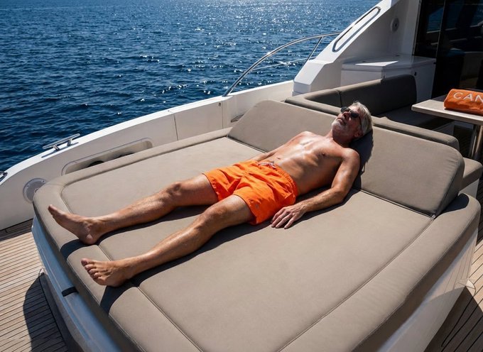 A shirtless man in bright orange swim trunks sunbaths on the cushioned deck of a luxury yacht on calm blue waters, with the partial text "CA" visible on an orange towel nearby. The image evokes the "economic sovereignty" and wealth preservation referenced in the post, visually suggesting a lifestyle of generational prosperity and leisure that Bitcoin advocates like Saylor promote as achievable through early adoption.