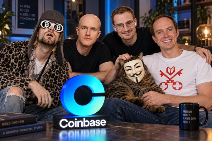 Group photo of four men including cryptocurrency personalities posing with a Coinbase logo display and what appears to be an Anonymous/V for Vendetta mask, with a person wearing '₿' (Bitcoin) symbol glasses; books and a 'FREEDOM MONEY A WIDE OPEN WORLD' mug are visible.
