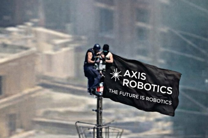 Two people stand atop a tall structure waving a black flag that reads 'AXIS ROBOTICS - THE FUTURE IS ROBOTICS' with a cityscape visible in the background.