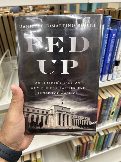 A hand holds up the book 'Fed Up: An Insider's Take on Why the Federal Reserve Is Bad for America' by Danielle DiMartino Booth in a library.