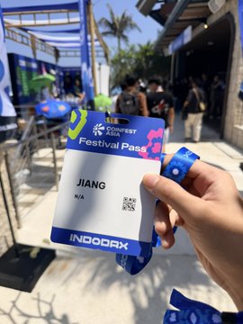 A hand holds up a 'CoinFest Asia' festival pass bearing the name 'JIANG' along with an INDODAX wristband, with an outdoor venue visible in the background.
