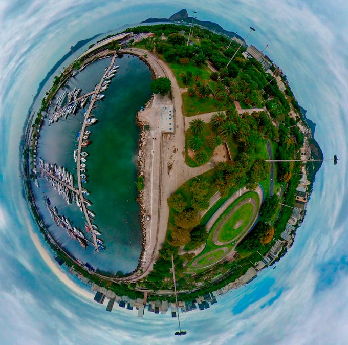 A 360-degree tiny planet panorama showing a coastal marina filled with docked boats, green parkland with palm trees, and a surrounding blue sky.