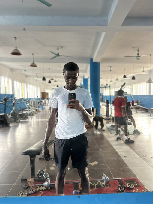 A young man in a white t-shirt and black shorts taking a gym selfie in a mirror while holding a dumbbell, with other people working out in the background.