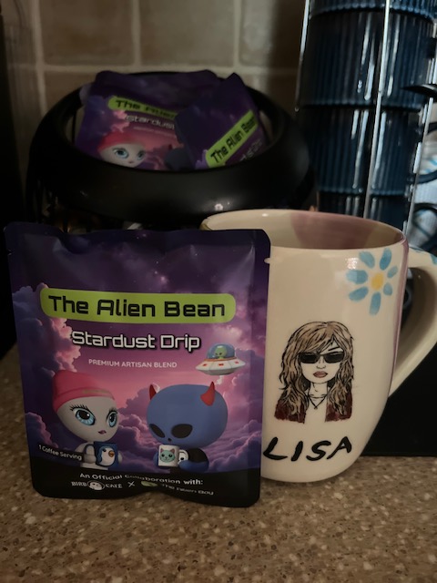 Two purple pouches labeled 'The Alien Bean Stardust Drip Premium Artisan Blend' coffee sit on a counter next to a beige mug decorated with a cartoon woman and the name 'LISA'.