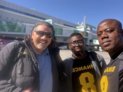 Three men smile for a selfie as they pose in front of a building, one wearing a shirt with "Binance" and "8" visible, celebrating the company's 9th anniversary.
