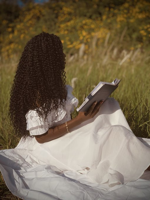 A woman with long curly hair wearing a white dress sits in a grassy field reading a book, with yellow flowers and tall grass in the background.