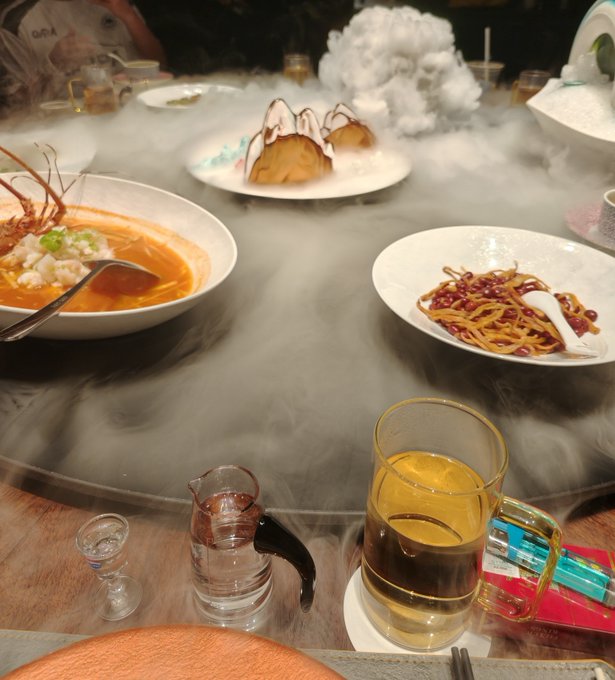 An elaborate dining experience featuring multiple dishes surrounded by a thick layer of dry ice fog, including an orange soup, a noodle dish with pomegranate seeds, and what appears to be a mushroom dessert, accompanied by tea and glassware on a wooden table.