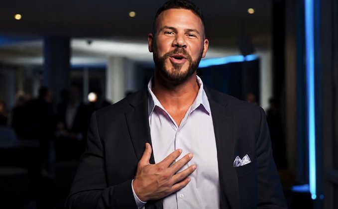 A man with a beard wearing a dark suit jacket over a light purple shirt, with a pocket square, gesturing with his hand over his chest in what appears to be an upscale venue.