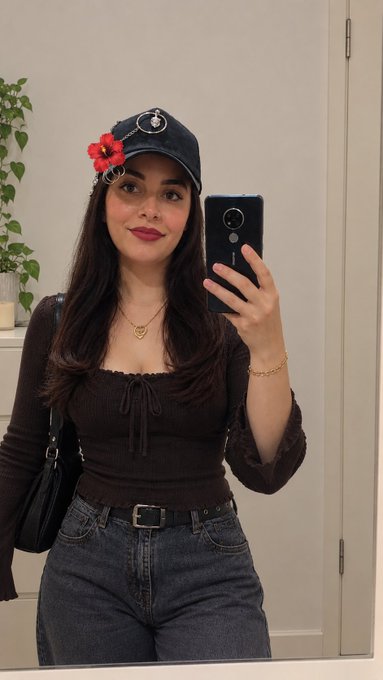 A mirror selfie of a young woman with dark hair wearing a black long-sleeved top with a front tie, blue jeans, a denim baseball cap adorned with red flowers, and a heart necklace.