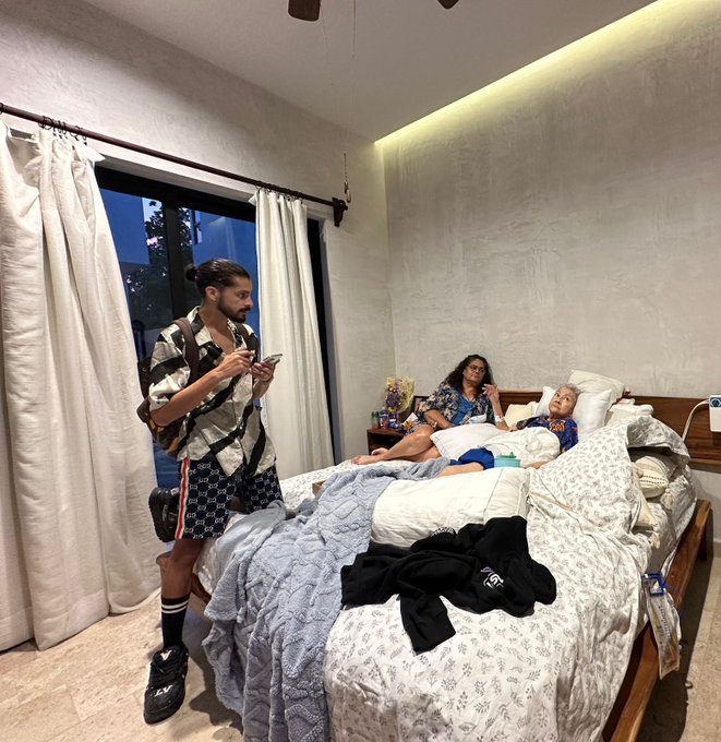 A family gathered in a bedroom; a man stands looking at his phone, while a woman and two children relax on a large bed with patterned bedding.