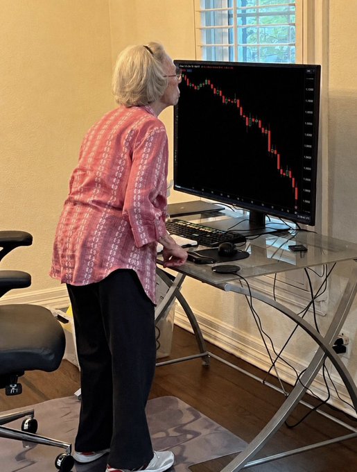 An elderly woman stands at a glass desk intently watching a steeply declining red candlestick chart on a large monitor, with an office chair beside her.