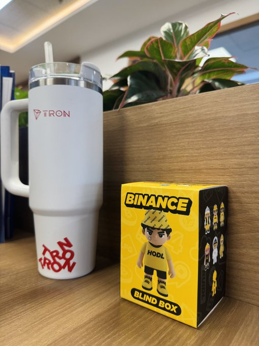 A white 'TRON' branded travel mug sitting next to a yellow 'BINANCE BLIND BOX' featuring a 'HODL' character figure on a wooden desk.