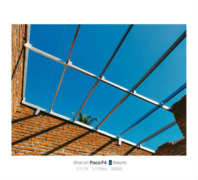 A photo taken on Poco F4 Xiaomi showing the underside of a brick wall roof structure with metal beams against a clear blue sky, with a palm tree visible in the distance.
