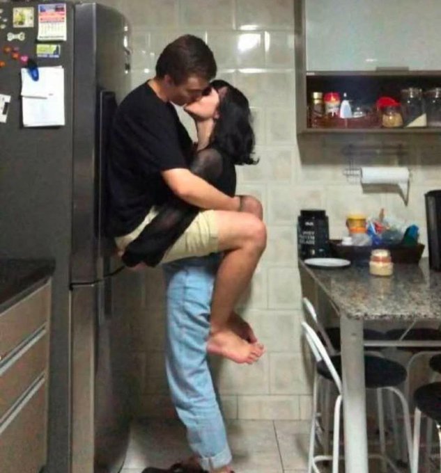 A couple shares a passionate kiss in a kitchen, with the man lifting the woman as she wraps her legs around him near a refrigerator and kitchen counter.
