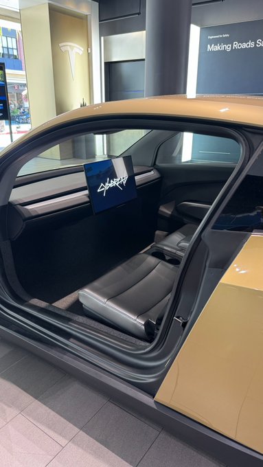 Interior photo of a yellow sports car (appears to be a Tesla Roadster) shown from the open door perspective with a signature on the dashboard screen, displayed in a Tesla showroom with 'Making Roads' text visible in background.