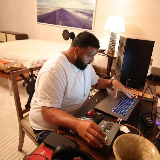 A bearded man in a white t-shirt sits at a hotel room desk working on a laptop with audio production equipment, headphones, and studio monitors around him.