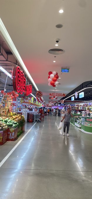 An interior photo of a supermarket aisle decorated with red Chinese New Year ornaments, balloons, and a 'NEW YEAR NEW LOOK' banner, with a shopper walking down the polished floor.