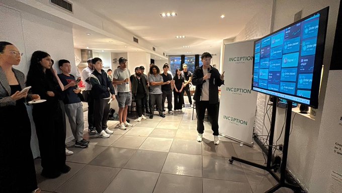 A photograph of an indoor presentation or networking event with a diverse crowd watching a speaker present information on a large screen displaying an 'INCEPTION' interface.