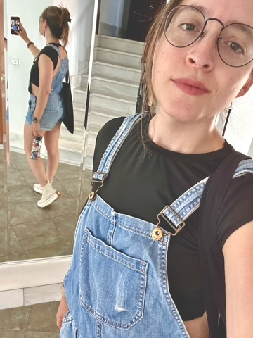 A selfie of a woman wearing round glasses and a black t-shirt under denim overalls, standing in front of a mirror that reflects her outfit.